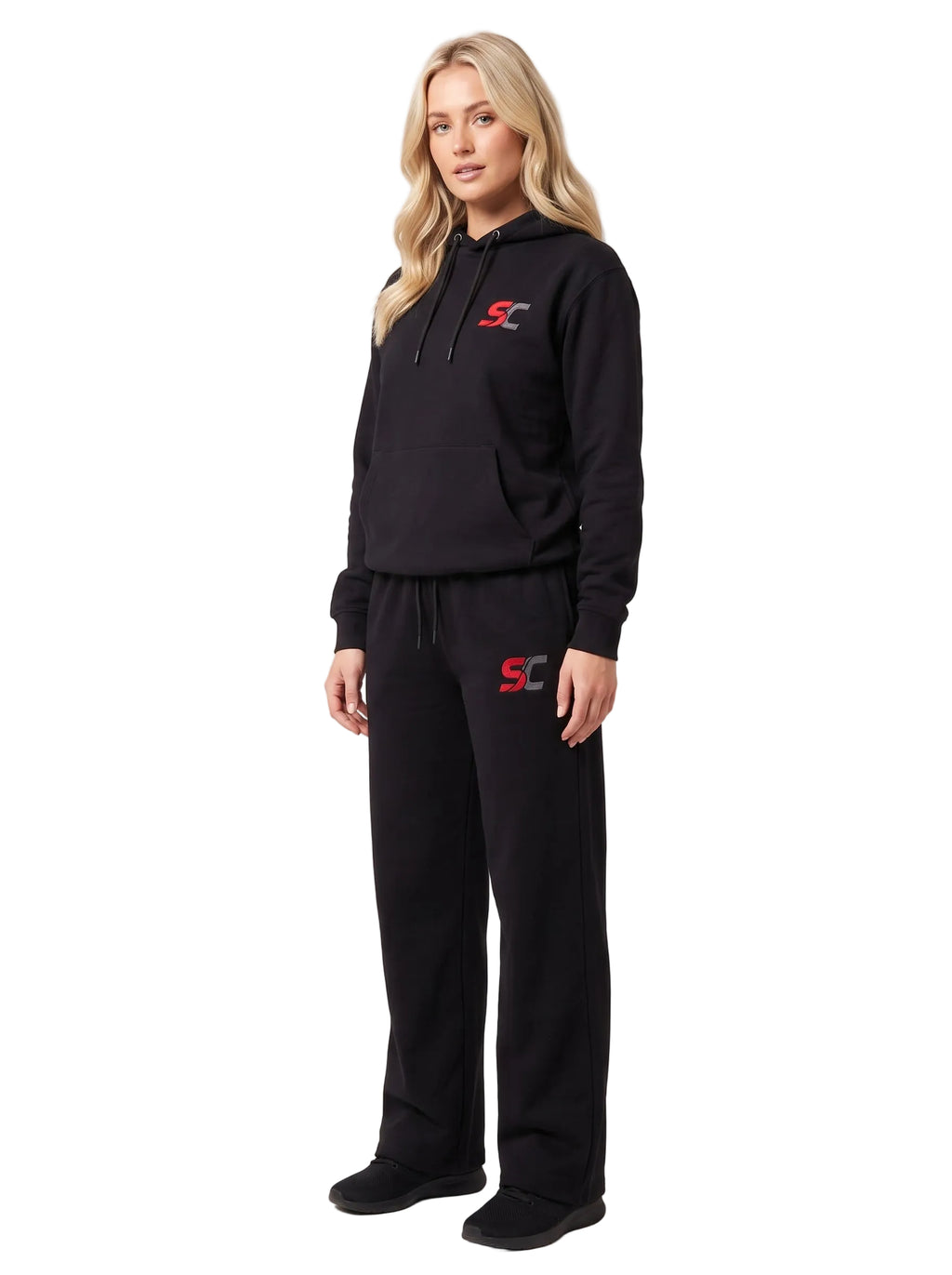 A woman modeling a black two-piece tracksuit with a pullover hoodie and matching sweatpants, featuring a red and white 'SC' logo on the chest and left thigh, set against a plain light gray studio background.