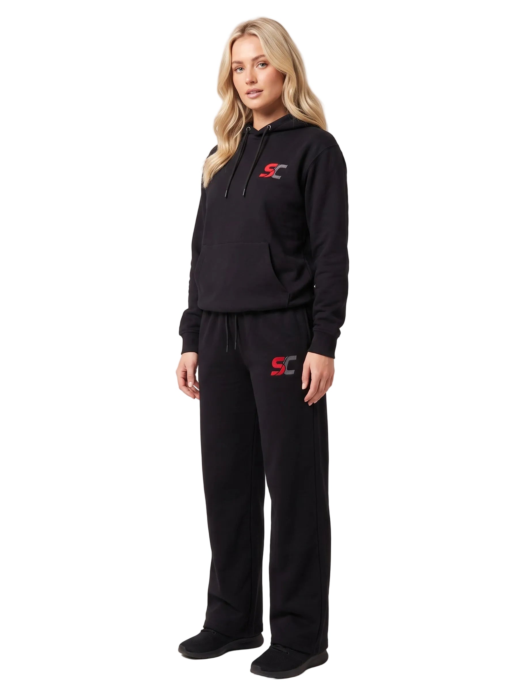 A woman modeling a black two-piece tracksuit with a pullover hoodie and matching sweatpants, featuring a red and white 'SC' logo on the chest and left thigh, set against a plain light gray studio background.